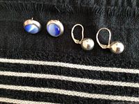 Photo of two pairs of silver earrings placed on black fabric: one pair of blue stone stud earrings and one pair of silver drop ball earrings