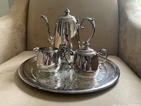 Photo of entire silver tea set on a round tray showing tea pot, creamer, sugar bowl, and tray.