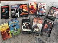 Photo of 11 VHS movie tapes arranged in rows on a surface showing front covers including Witness, Lethal Weapon, Armageddon, Spider-Man, My Baby's Daddy, The Fugitive, What Dreams May Come, The Sound of Music, Apollo 13, Titanic.