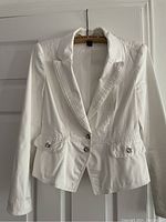 Front view of white women's blazer with silver buttons and flap pockets hanging on a wooden hanger.
