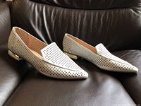 Side angle view of the pair of white leather women's flats with perforated design and gold detail on heel, placed on leather sofa.