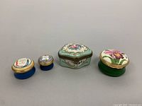 Photo of four assorted small decorative pill boxes including antique porcelain and Halcyon Days enamels arranged in a row.