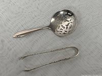 Photo showing the pierced serving spoon and sugar tongs side by side on a fabric surface.