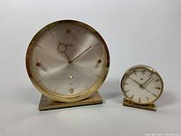 Two brass clocks side by side: a larger Swiza calendar clock and a smaller Ames travel alarm clock, both with silver faces.