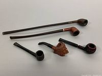 Five assorted vintage tobacco pipes displayed on a white background, showing variety in stem length and design including long Piccadilly and Churchwarden types.