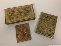 Three brass items: one rectangular brass box with detailed top engraving, one brass plaque with King Arthur and the Round Table scene, one brass bas-relief plaque of St George on horseback.