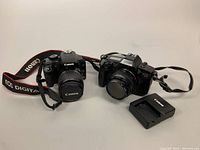 Canon EOS Rebel XS digital camera with 18-55mm lens, Canon EOS 650 35mm film camera with 50mm lens, and Canon battery charger shown together on table.