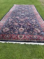 Full view of large Turkish wool carpet with red border and navy blue floral field laid on grass.