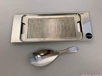 Top view of the rectangular stainless steel fish/vegetable grate and broad risotto spoon, polished finish, Alessi brand sticker visible on grate.