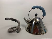 Side view of silver-plated spiral tapered candleholder (Dansk) and polished aluminum kettle with blue handle (Alessi) on white background