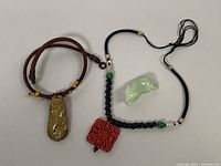 Full shot of the three pendants with cords laid flat: cinnabar square pendant on black cord, jade tiger pendant, jade gourd pendant on brown cord.