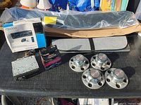 Overview of automotive lot showing Clarion stereo in box, wireless alarm box, bundle of weather stripping, four chrome wheel center caps, and smaller automotive parts.