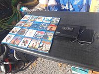 Black LG Blu-ray player with remote control on top, black external drive, and Blu-ray movie cases on a black surface outdoors.
