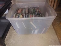 Clear plastic storage bin containing DVDs stored upright inside, showing full bin and partial view of DVDs