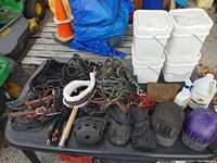 Overview of horse tack items including bridles, reins, bits, ropes, mounting block, horse grooming tools, and horse care liquids on a black plastic surface outdoors.