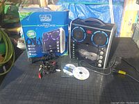 Portable karaoke machine with dual speakers, front control panel, microphone holder, cables, two karaoke discs, and original box with branding.