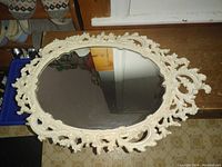 Front view of an oval mirror with an ornate, white painted decorative frame featuring floral and scroll motifs, reflecting part of the room.