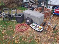 Photo showing all outdoor items including hose reels, storage box, hoses, fan, heater, small item tray, and string trimmer on ground covered with fallen leaves.