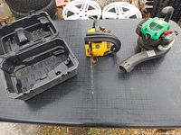 Mcculloch yellow chainsaw, green and black weedeater blower, and black case laid out on black table outdoors with tires and wheels in background