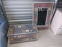 Photo showing a small rectangular wooden chest with colorful tile inlays on top and front, and a matching large rectangular mirror with a frame featuring the same tile inlay patterns, both set on the floor in a storage area.