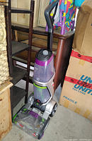 Electrolux upright steam cleaner showing handle, water tank, and cleaning base in storage surrounded by wooden shelving and cardboard boxes.