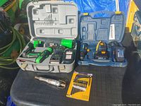 Overview of lot with green cordless drill, Mastercraft tools case, pneumatic air tool, and drill bit package on black surface outdoors