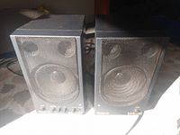 Pair of vintage black Roland MA-110 bookshelf speakers with metal mesh grille on front showing woofer and tweeter drivers, control knobs and power switch visible on front panel, slightly dusty.