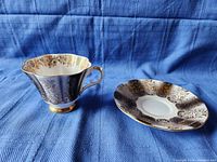 Teacup and saucer set placed on blue fabric background, showing front and side view highlighting gold floral decoration and scalloped edges.