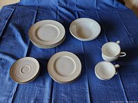 Photo showing stacked large plates, medium plates, two cups with handles, and saucers on a blue cloth background.