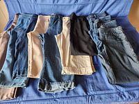 Overview of petite women's capris, jeans, and dress pants laid out on blue fabric background. Six capris, four jeans and one black dress pants visible.
