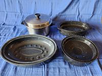 Four pieces including three roasting pans of different sizes and one round pot with lid, all with speckled black enamel finish.