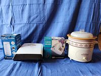 Photo shows four small kitchen appliances against a blue cloth backdrop: electric coffee grinder in box, George Foreman grill, hand mixer in box, crock pot with floral pattern and glass lid.