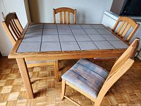 Rectangular dining table with wood frame and ceramic tile top, surrounded by four wooden chairs with worn fabric padded seats.