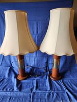 Full view of pair of teak lamp bases with attached cream-colored fabric lamp shades featuring scalloped edges curling slightly at bottom. Both lamps are plugged in with cords resting on blue fabric background.