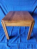 Pair of wooden side tables against a blue fabric background, showing a side view of one table clearly displaying the legs and smooth rectangular top with rounded corners.