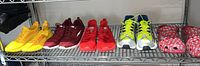 Five pairs of size 11 shoes lined up on a metal shelf: bright yellow Aefe sneakers, maroon Adidas sneakers, red neon Adidas sneakers, silver and neon yellow Adidas sneakers, and red Vineyard Vines Crocs with pattern.
