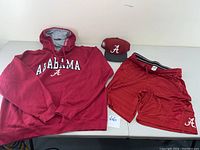 Photo showing all three items in the set: red Alabama hoodie, red Champion shorts, and red Alabama fitted hat laid out on a table.