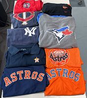 Photo showing folded MLB shirts featuring Baltimore Orioles, Minnesota Twins, New York Yankees, Toronto Blue Jays, and Houston Astros in various colors and sizes.