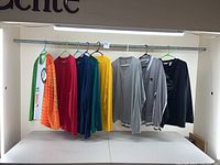 Seven athletic wear tops hanging on a metal bar: colorful Guinness Irish 5K shirts, solid thin long sleeves in red, blue, yellow, gray Champion long sleeve, black Reebok hoodie pullover.