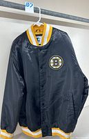 Front view of black nylon snap front jacket with Boston Bruins logo on chest and yellow and white striped collar, cuffs, and waistband.