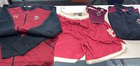 Full lot overview showing Under Armour Boston College Eagles zip jacket, maroon shorts with white and gold trim, maroon visor, and black/maroon polo shirt on white table
