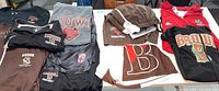 Wide view showing multiple Brown University hoodies, t-shirts, letterman style jacket, shorts, and other apparel on table