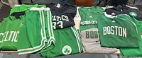 Multiple Boston Celtics apparel items including shorts, sleeveless jersey, t-shirts, and pants laid out on table