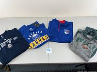 Four hockey apparel items laid side by side: Winnipeg Jets long sleeve shirt, Buffalo Sabres sweatshirt, New York Rangers quarter zip pullover, Minnesota Wild T-shirt.