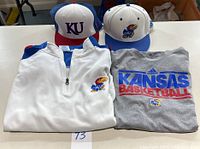 Two fitted hats in red, blue and white, a white quarter-zip pullover, and a grey Kansas Basketball t-shirt displayed together.
