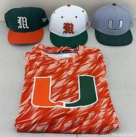 Three Miami Hurricanes fitted hats (green/orange, white/green, gray/green) and one orange Miami Hurricanes t-shirt laid out flat.