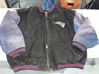 Full view of the New England Patriots black suede jacket with blue sleeves and red-blue striped trim on collar, cuffs, and waistband. Jacket shows signs of wear and discoloration.