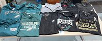 Lot of nine Philadelphia Eagles apparel items laid flat on a table showing various Nike and Adidas branded jackets, shirts, and polos in green, black, and grey with team logos and text.