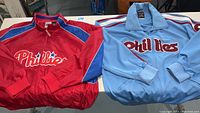 Two Phillies jackets laid flat on a table: red HomeBase Collection and light blue Cooperstown Collection.