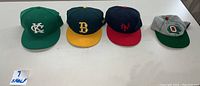 Four retro MLB caps displayed in a row: Kansas City (green with white logo), Boston (navy blue with yellow brim and yellow B logo), New York (navy blue with red brim and red NY logo), Oakland (grey with green brim and red O logo).